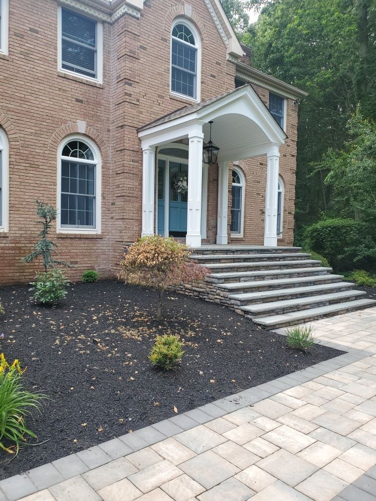 Brick house entrance with white columns, steps, and a stone walkway beside landscaped mulch beds.