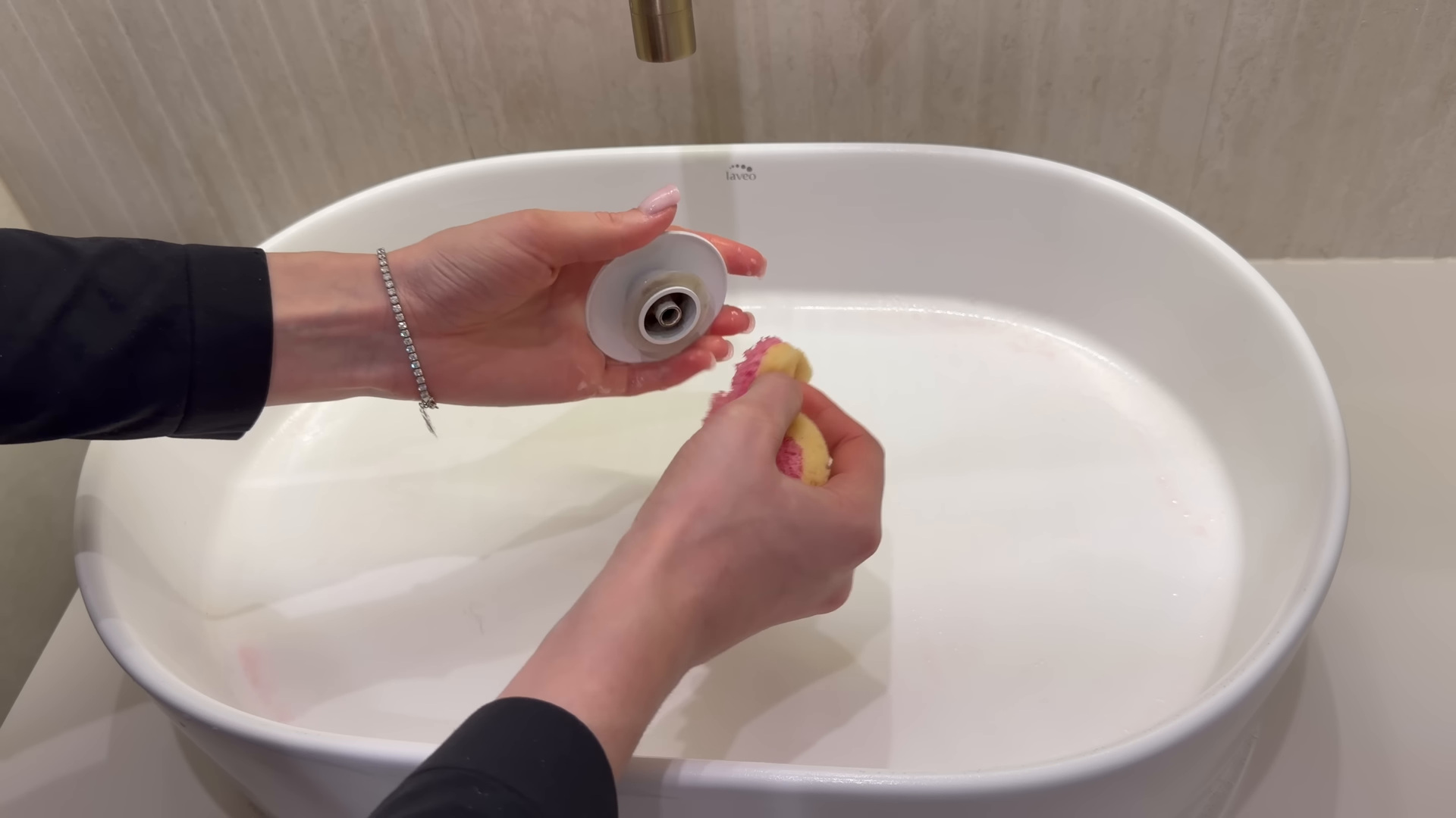 Hands rinsing a small gray device over a white basin with running water.