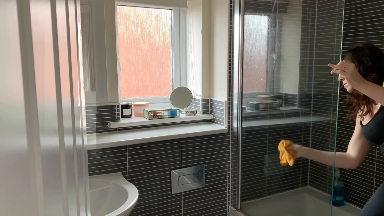 Person cleaning a tiled bathroom shower with a yellow cloth next to a frosted glass door.