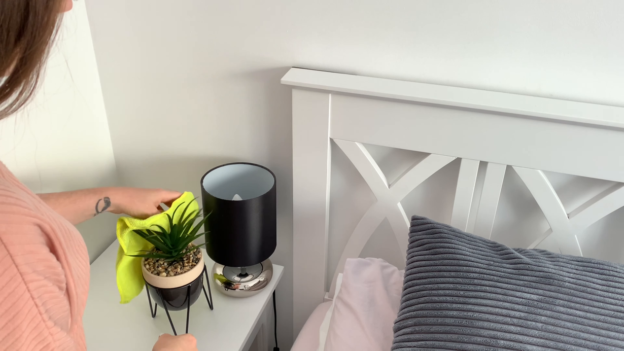 Hand placing a yellow cloth beside a small plant and black lamp on a bedside table next to a white headboard.