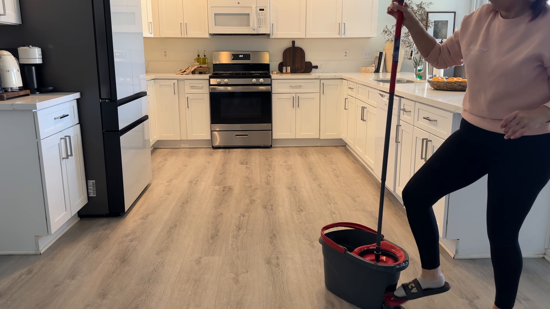 Person mopping a light wood kitchen floor with a red-and-black bucket nearby.