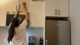 Woman reaching up to adjust a cabinet door in a kitchen beside a stainless-steel refrigerator.