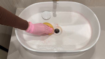 Hand wearing pink glove cleaning a sink drain in a white basin with yellow sponge