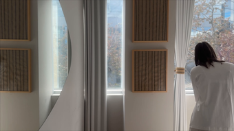 Person in a white shirt stands by a sunlit window with sheer curtains and woven wall panels.