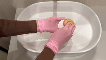 Gloved hands scrubbing a sponge in a white sink with soapy water