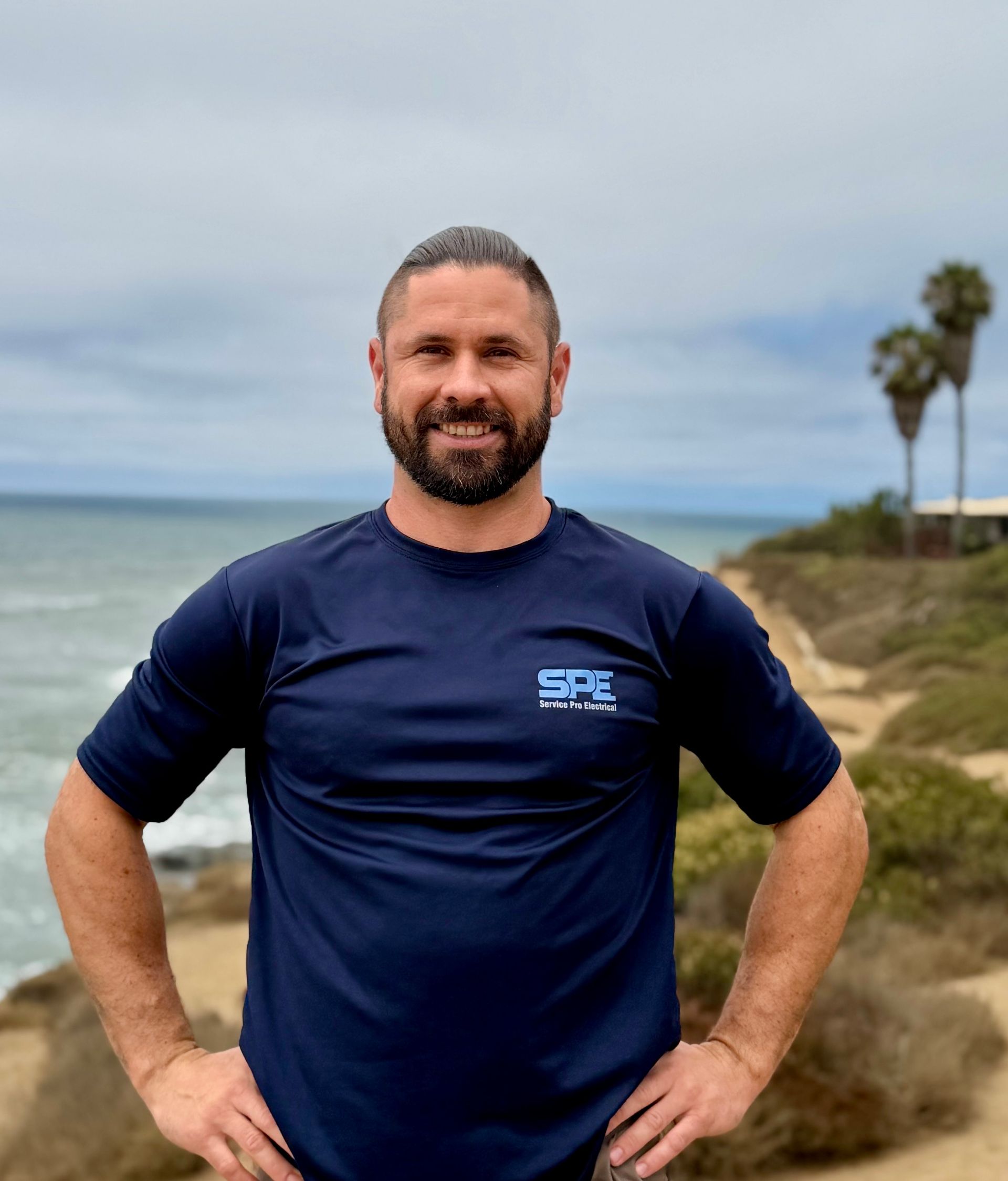 A man with a beard is standing in front of the ocean.