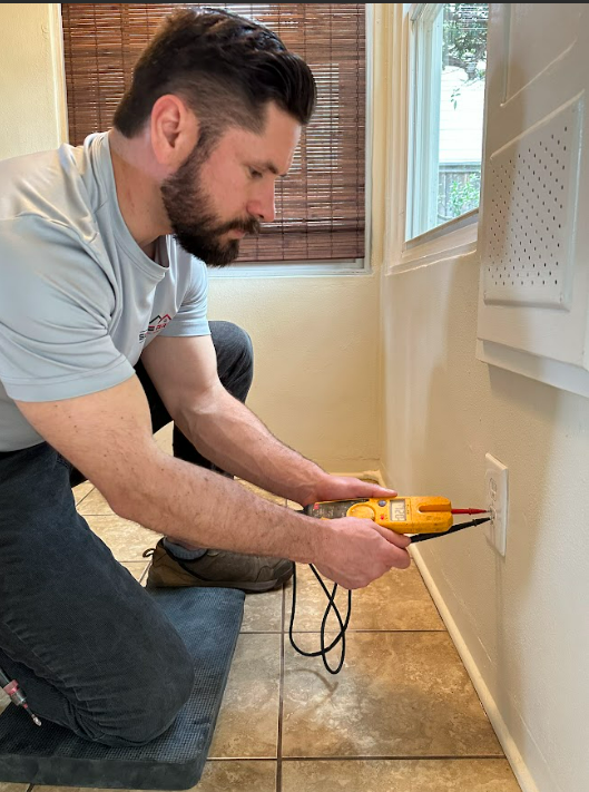 A man is kneeling on the floor using a voltmeter on a wall.