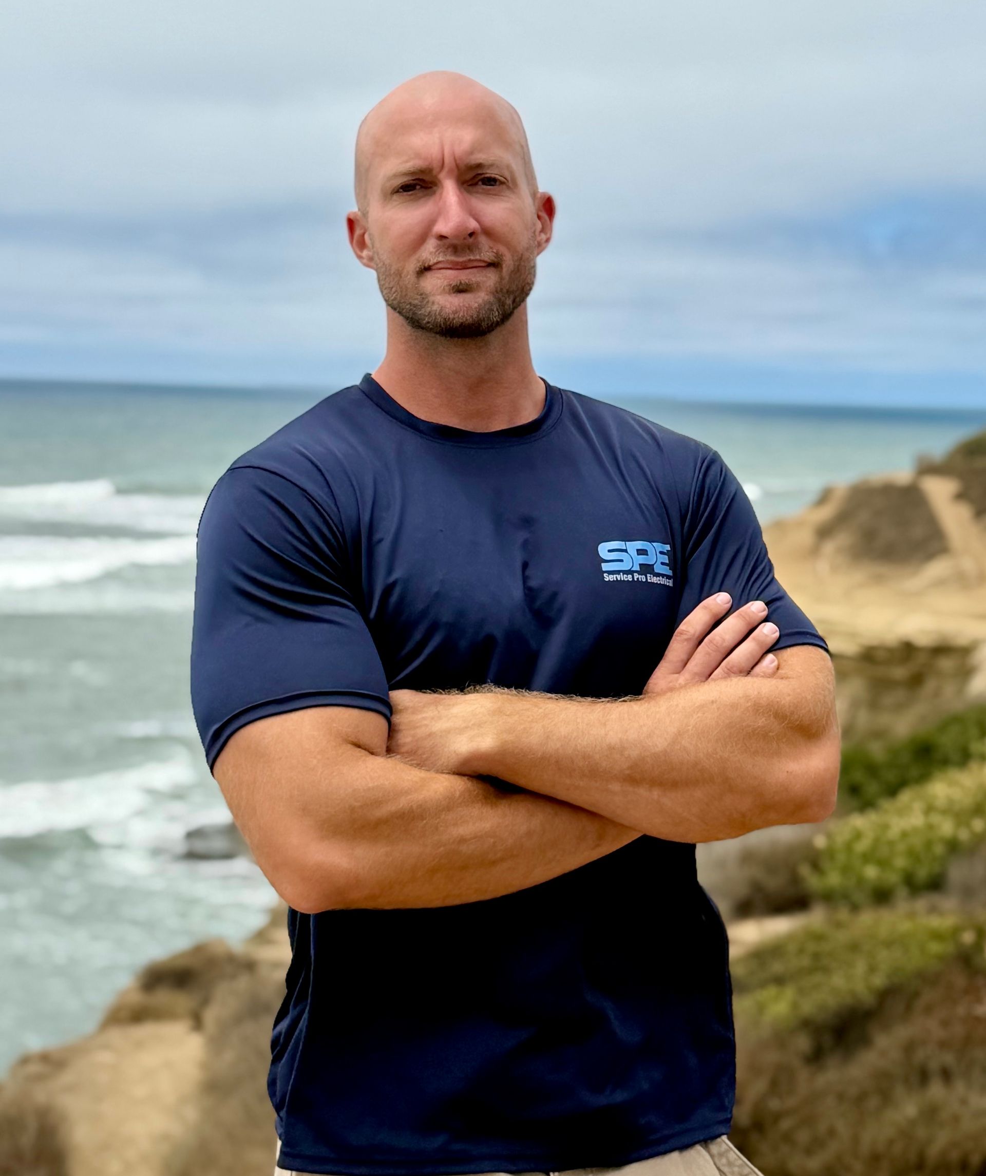 A bald man with a beard is standing in front of the ocean.
