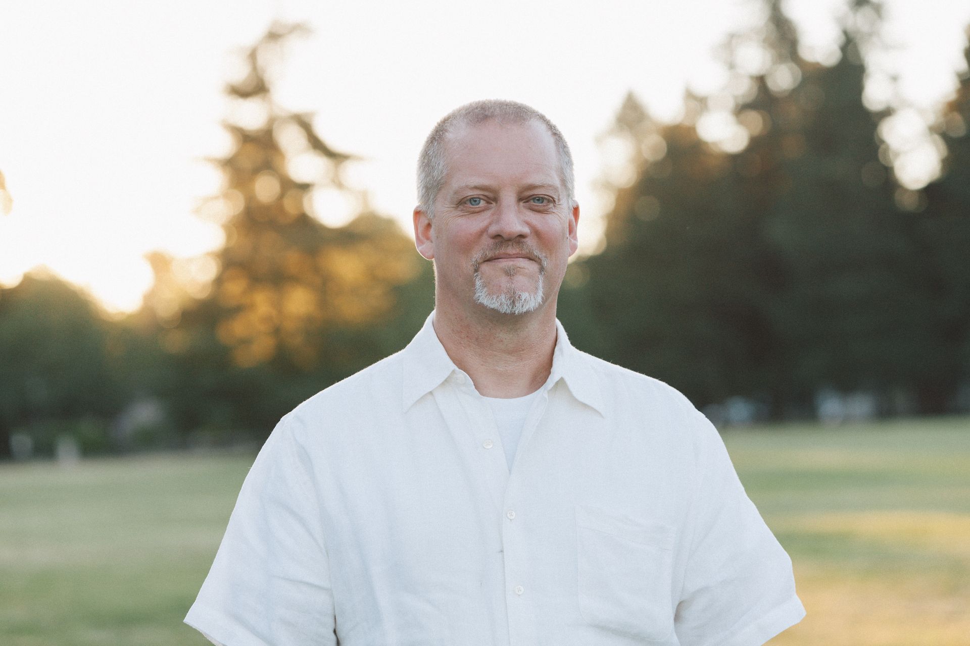 Man with graying beard and white shirt smiles outdoors in front of trees.