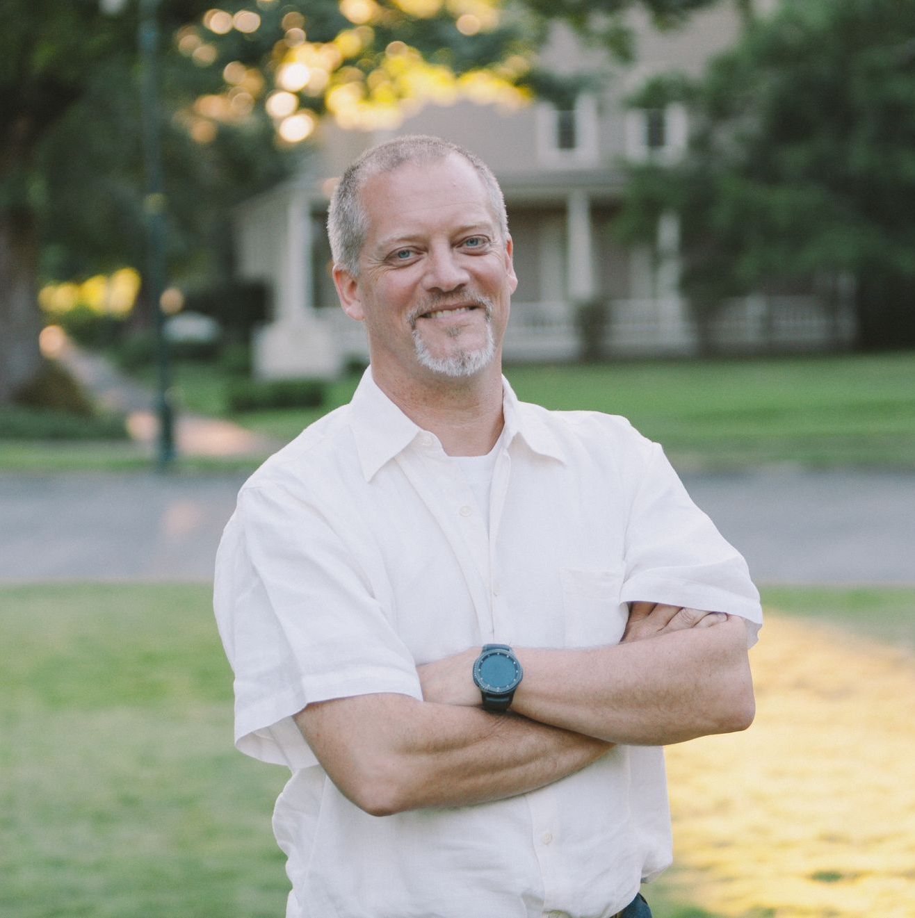 Man with graying hair and arms crossed smiles in front of a house.
