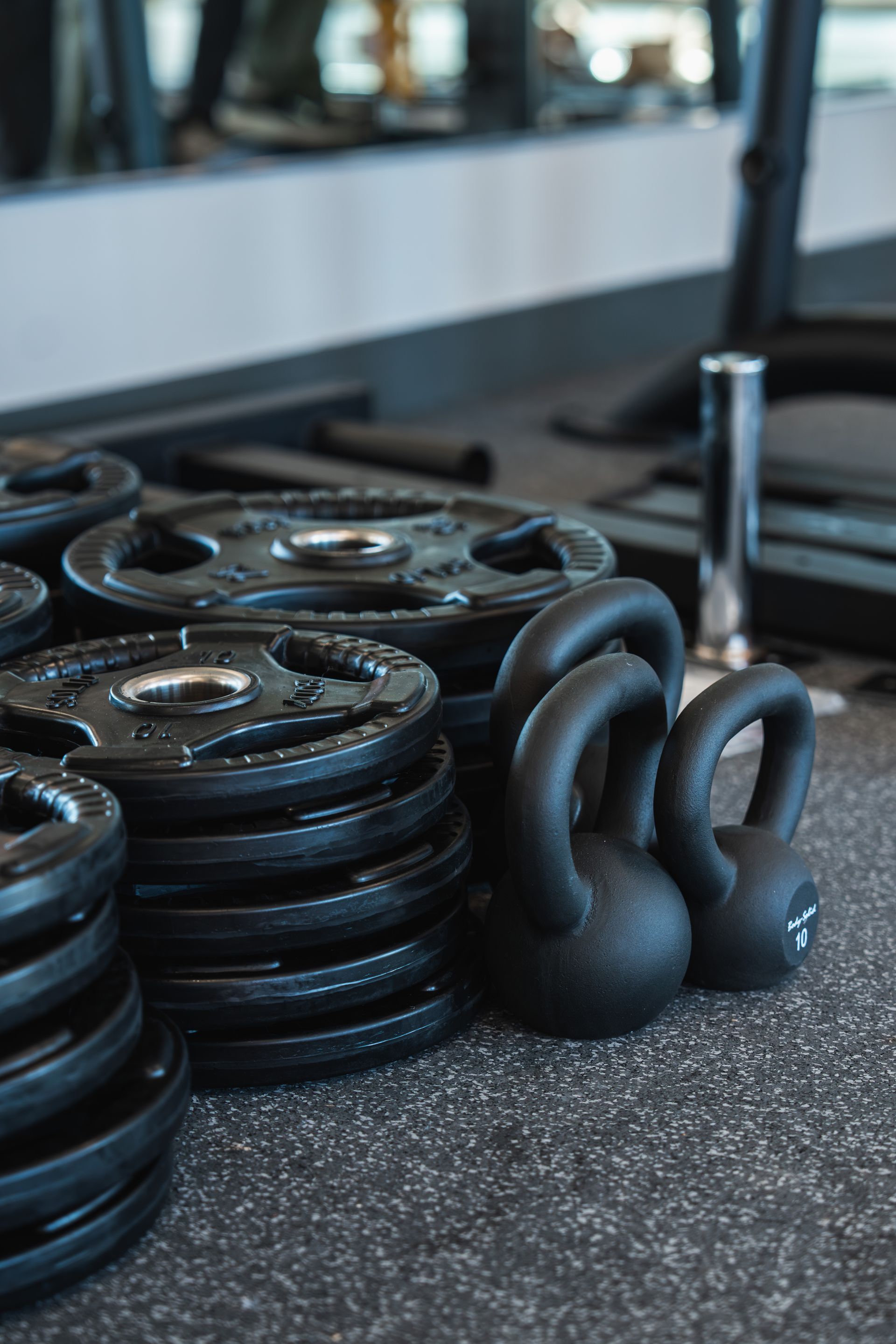 Black kettlebells and weight plates on a gym floor near a bench and mirror