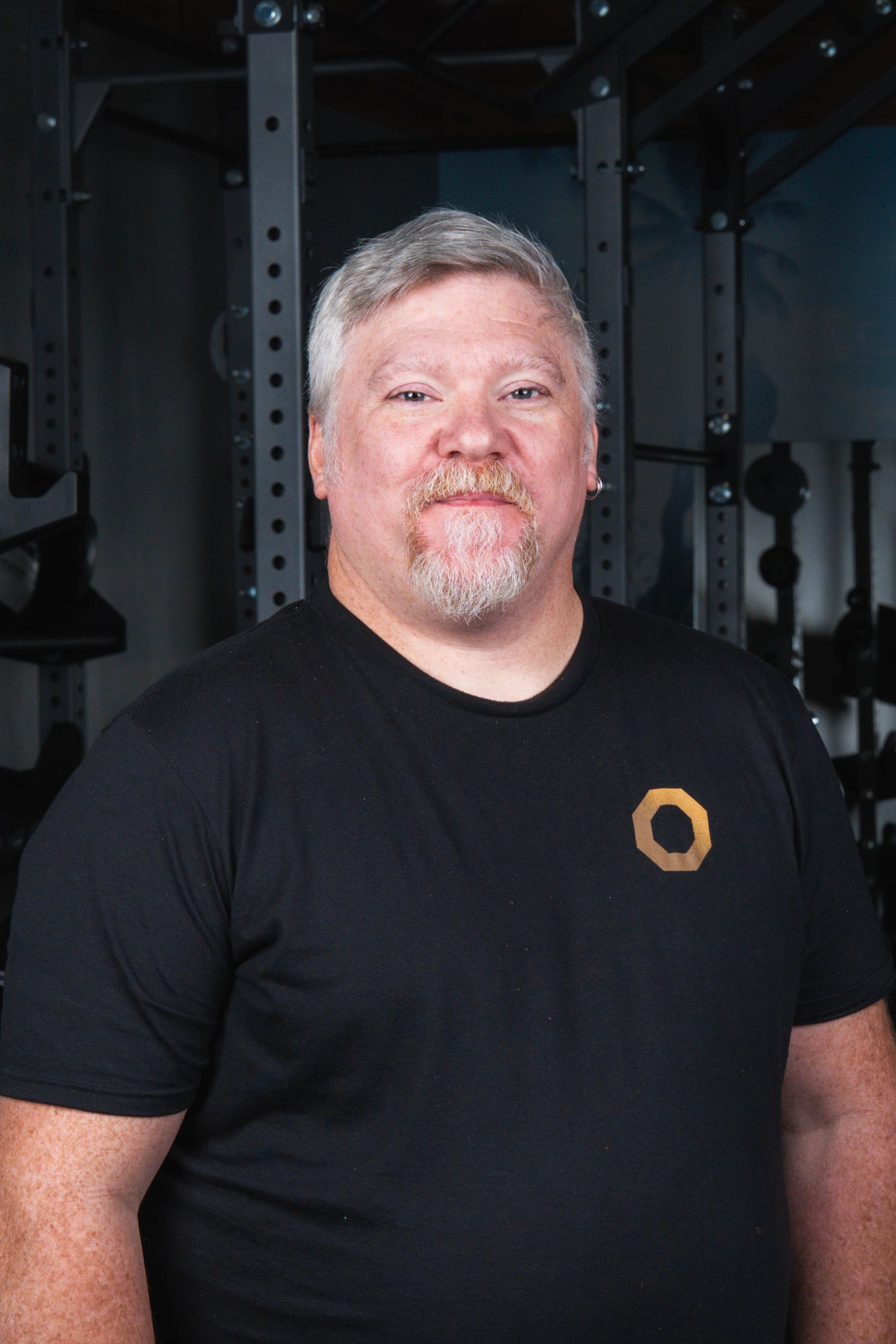 Man in black t-shirt with logo in a gym, smiling at the camera, weight racks in the background.