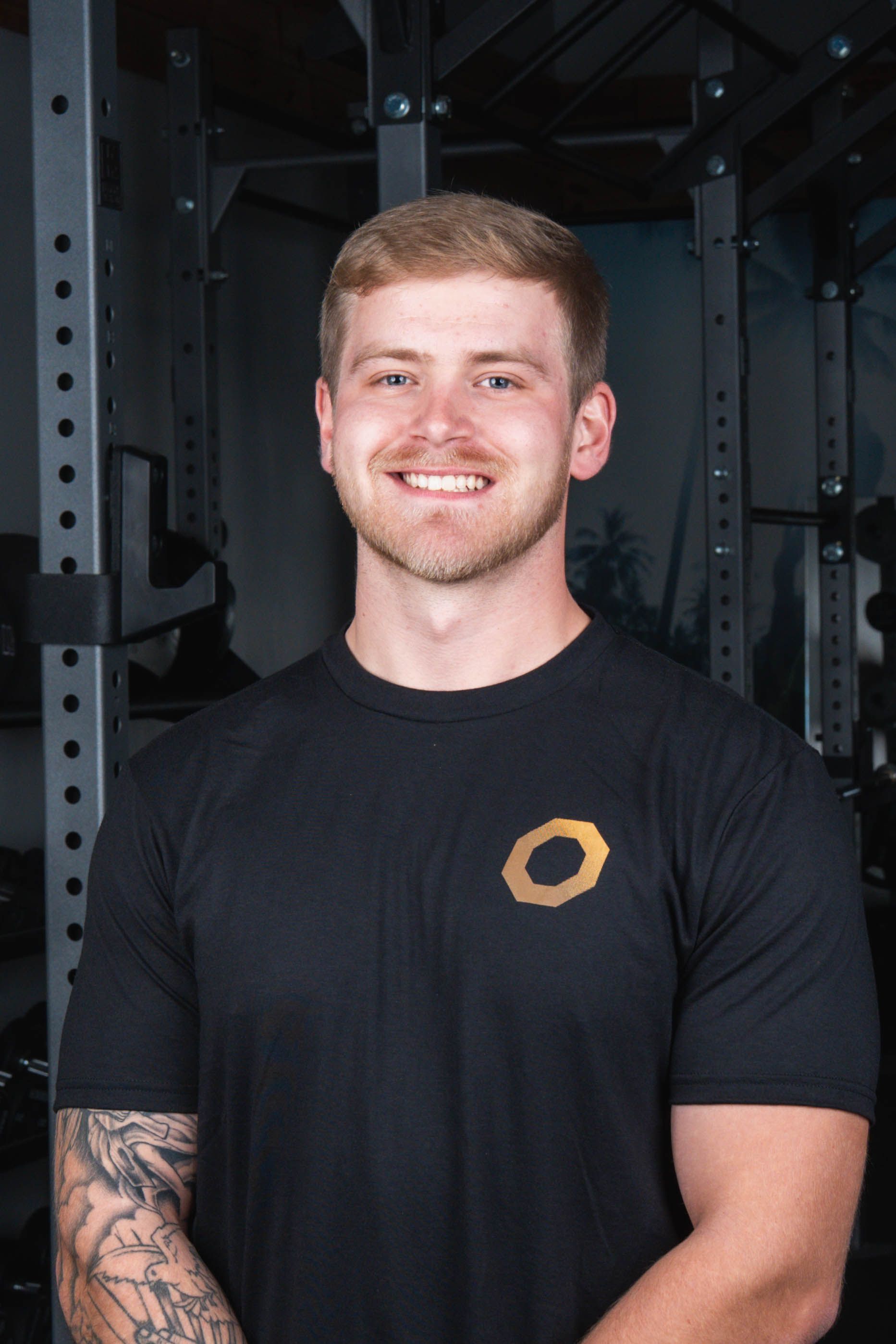 Smiling man in black shirt with logo, arms crossed, in a gym with weight racks.