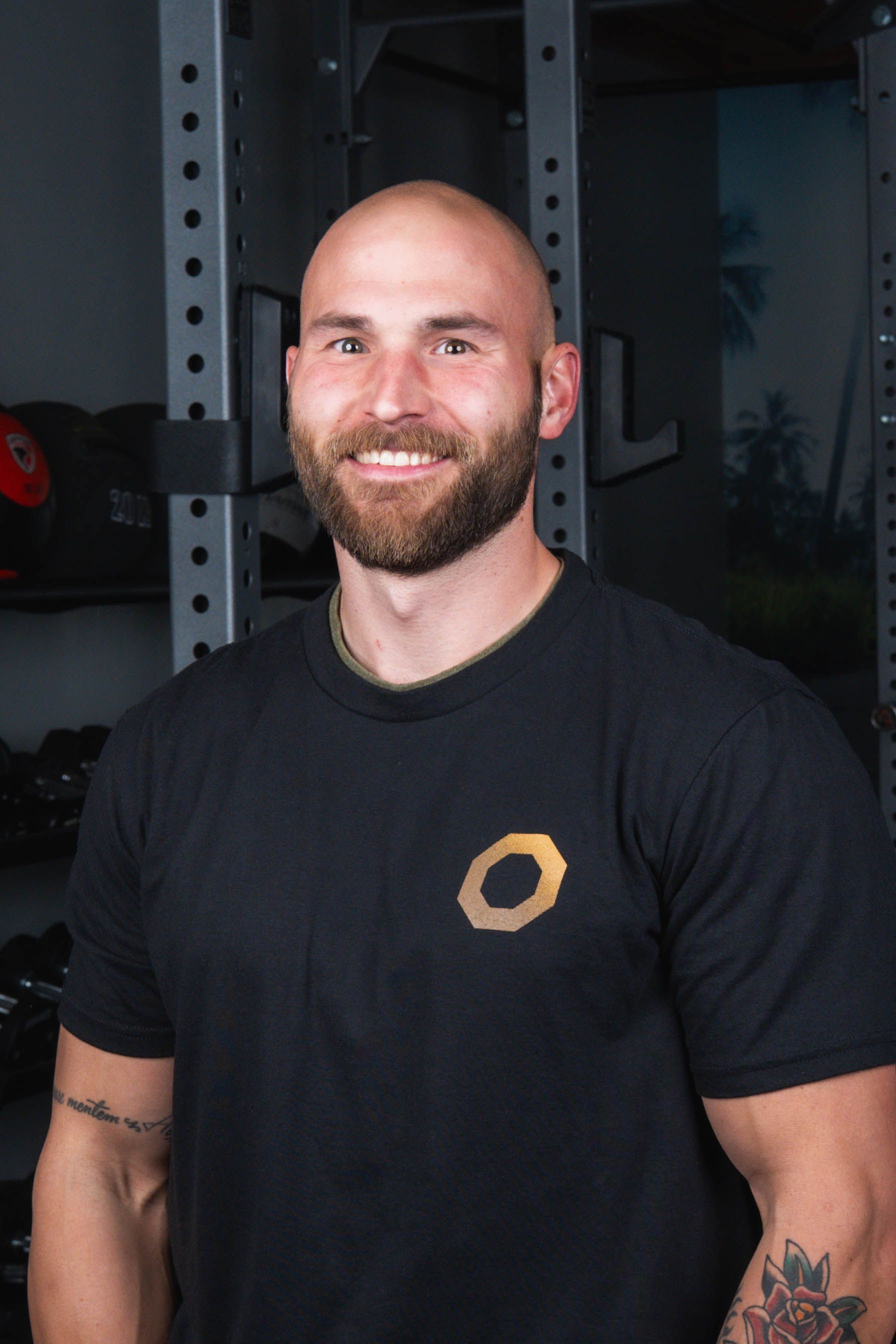 Bald man with beard smiles in a gym, wearing black shirt with logo.