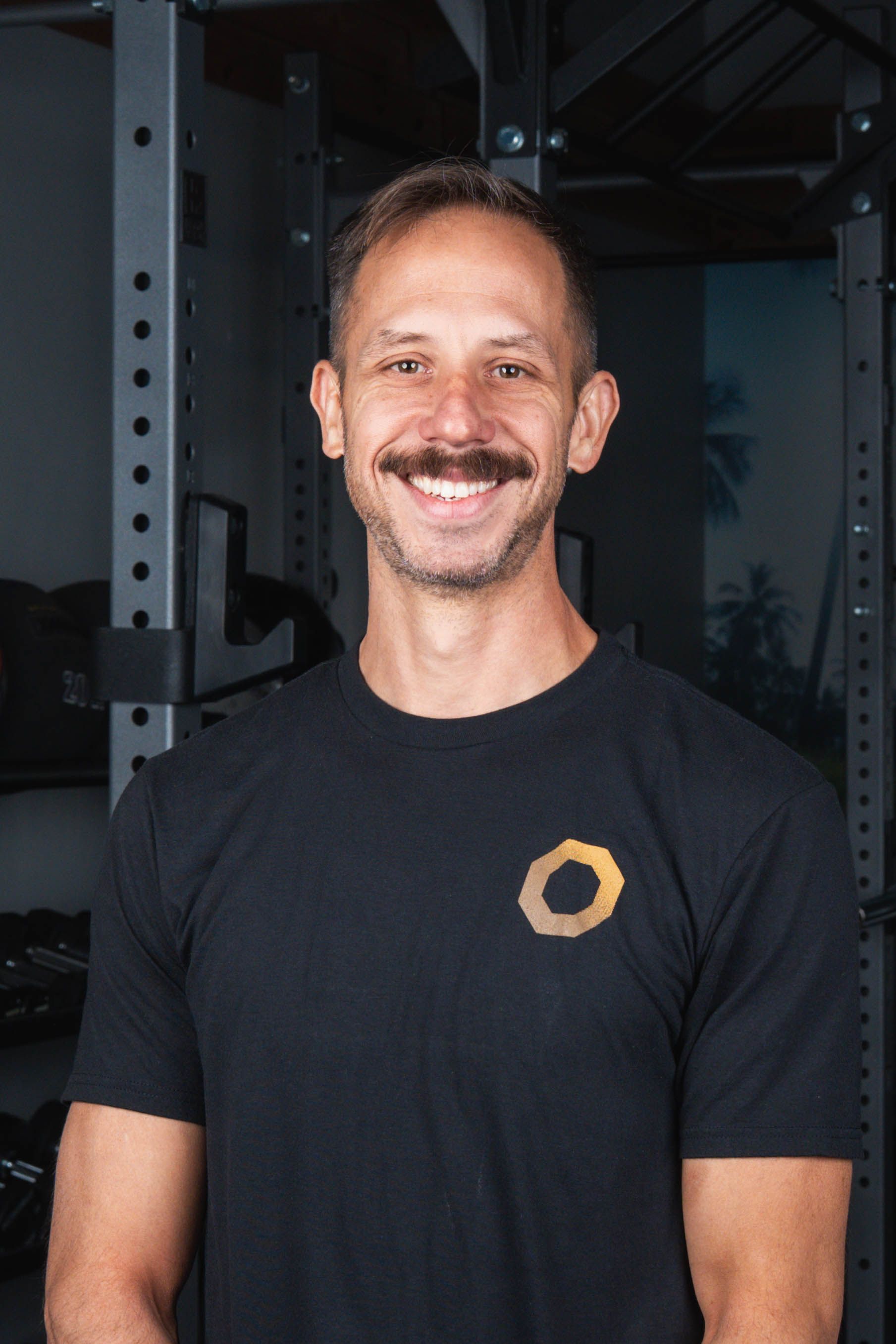 Man in black t-shirt smiles at the camera in a gym setting, with weight equipment in the background.
