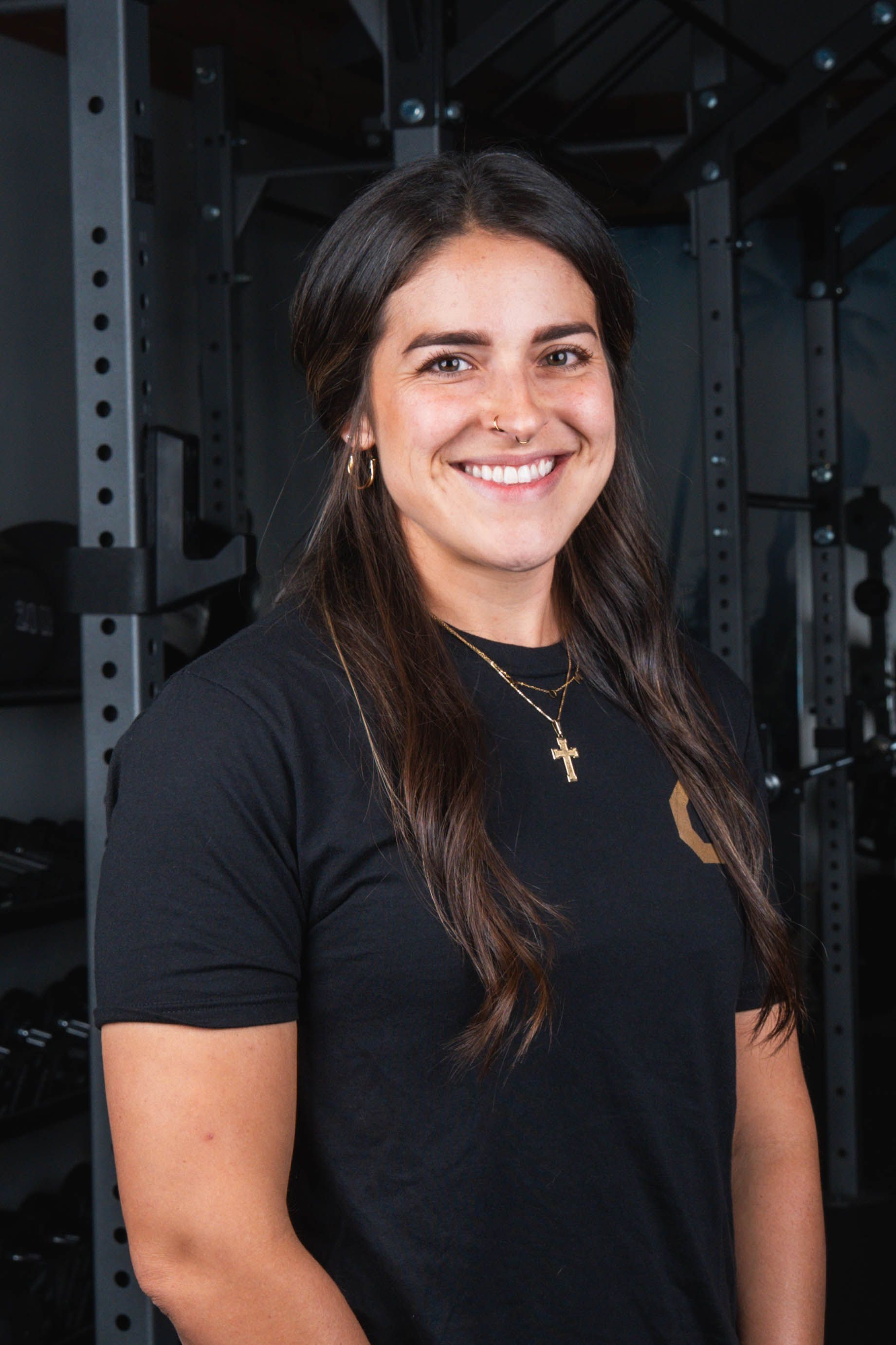 Woman smiling in a gym, wearing a black shirt with gold cross pendant.