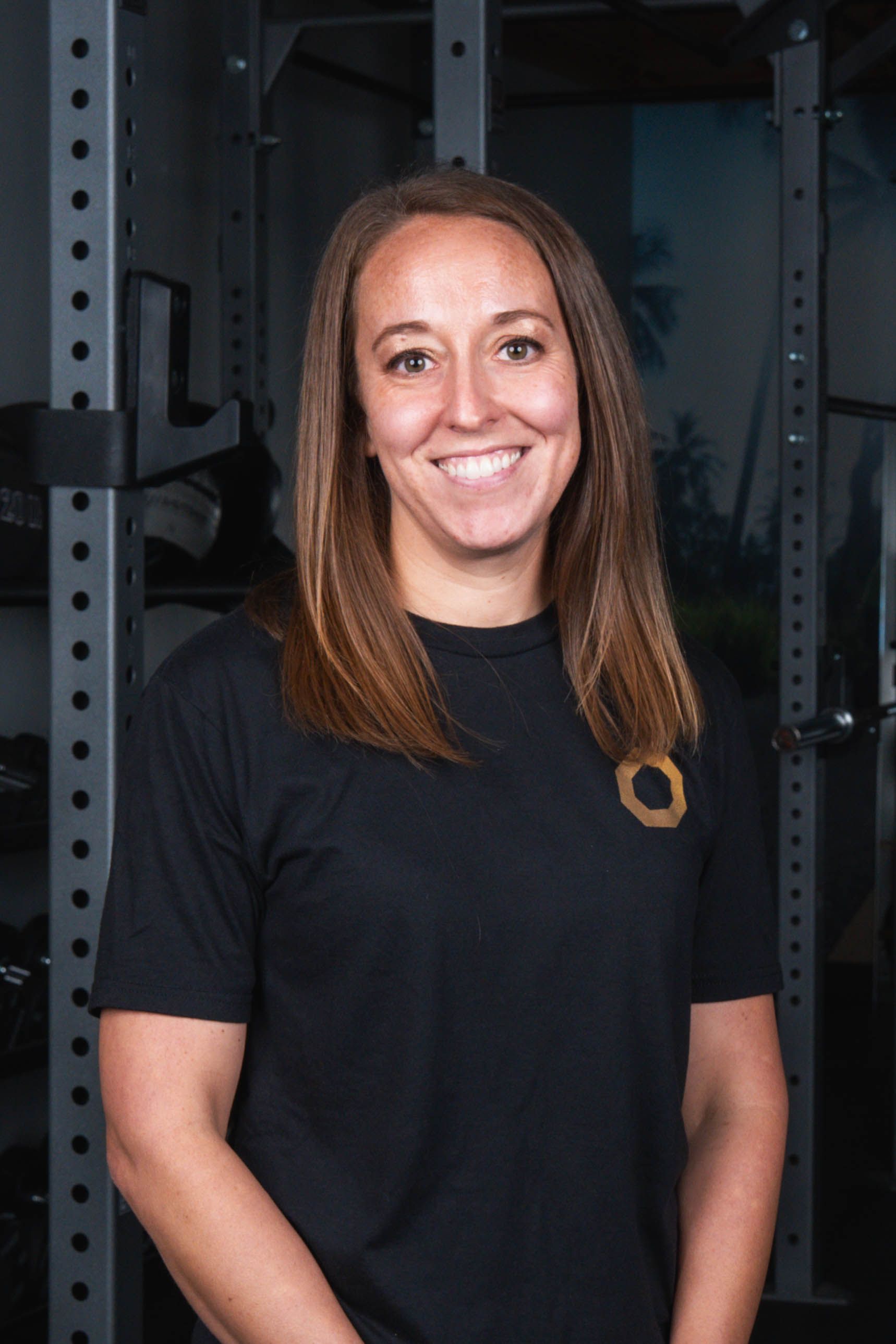 Woman smiling, standing in a gym. She wears a black shirt, leaning slightly forward, arms crossed.