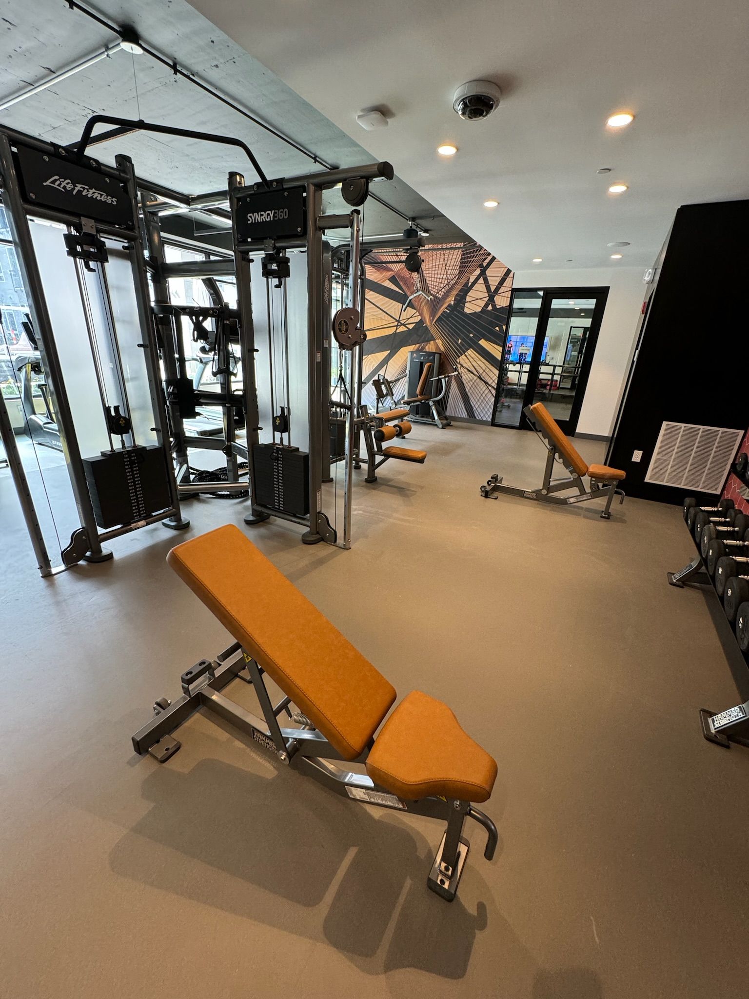 Bright gym with weight machine, orange incline bench, and mirrors along the wall