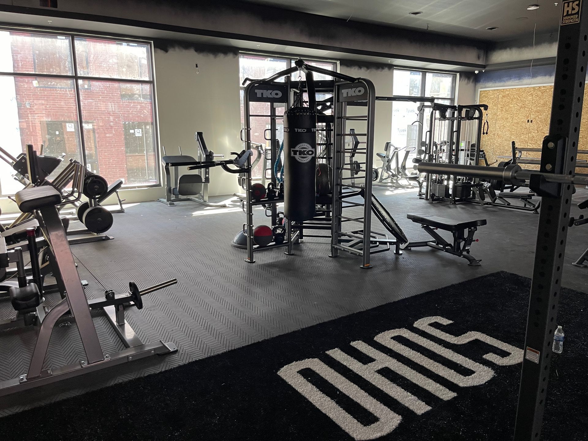 Modern gym interior with exercise machines, black flooring, and large windows letting in daylight.