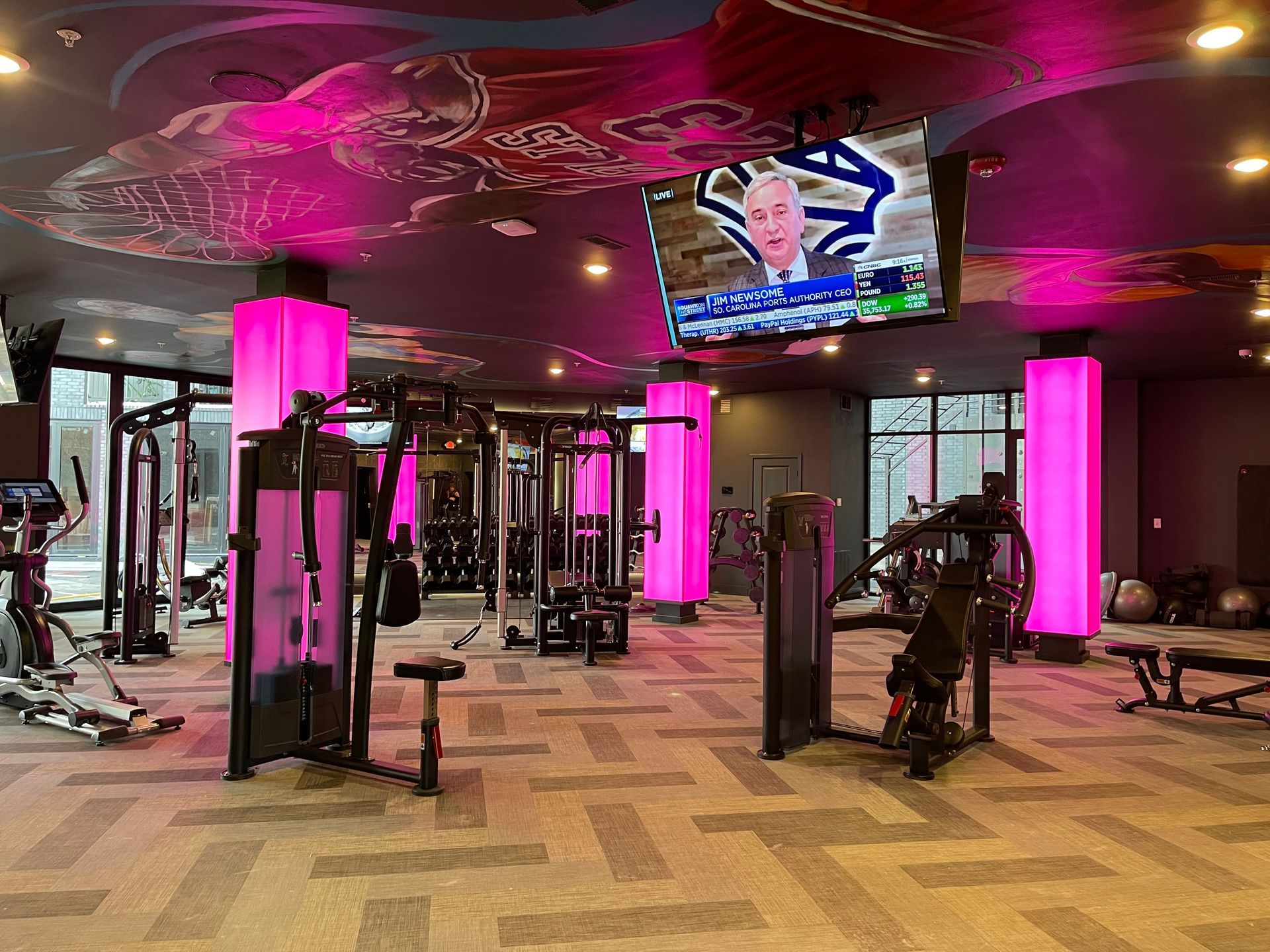 Modern gym with exercise machines, lit columns, and a TV showing a news broadcast.