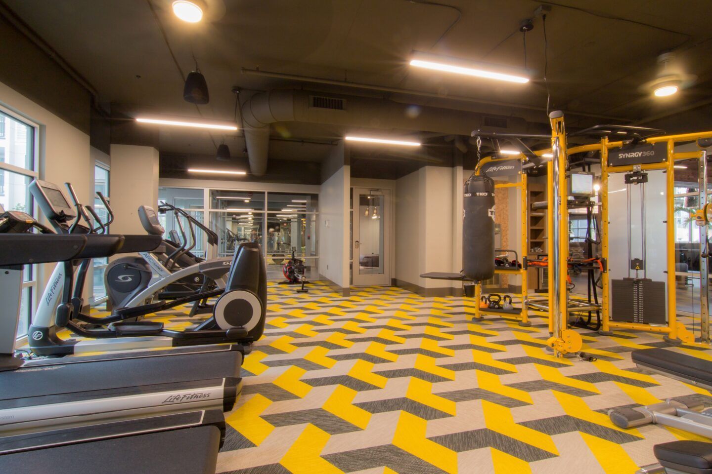 Gym with cardio machines, weight equipment, and a patterned yellow and gray floor.