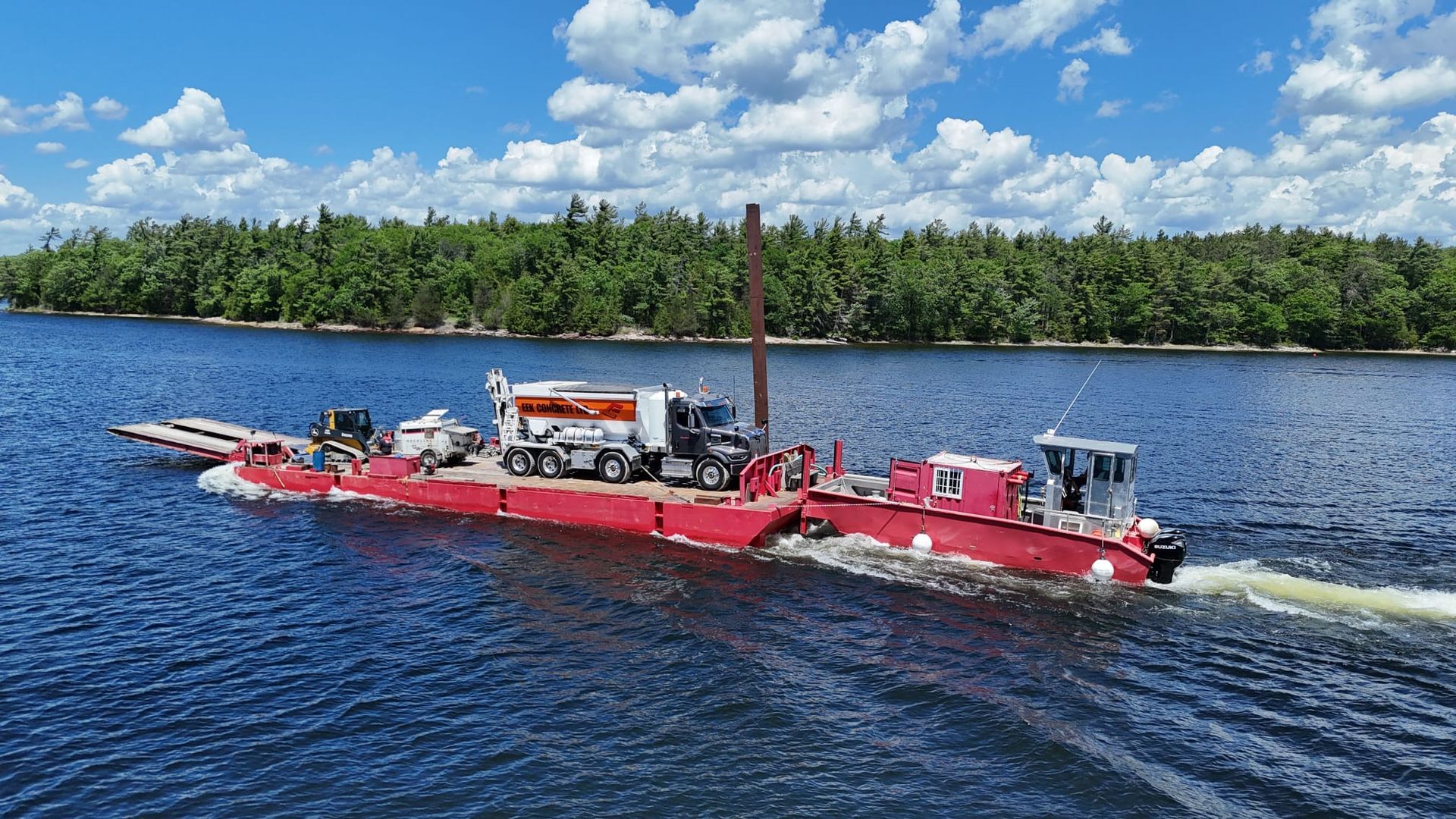 Red tugboat towing a long red barge across a blue lake with forested shoreline