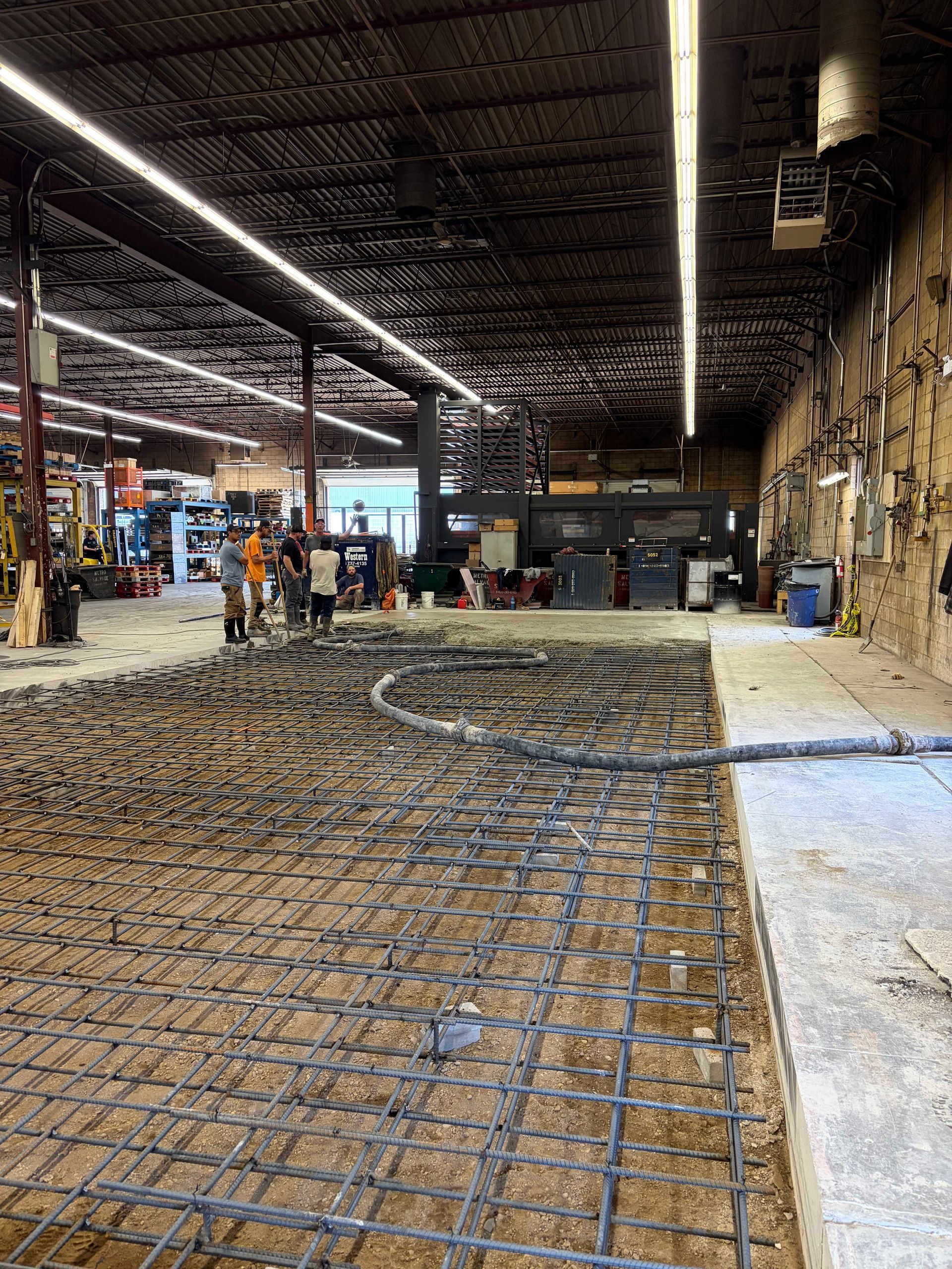 Concrete slab under construction inside a large industrial warehouse, with steel rebar grid in foreground.