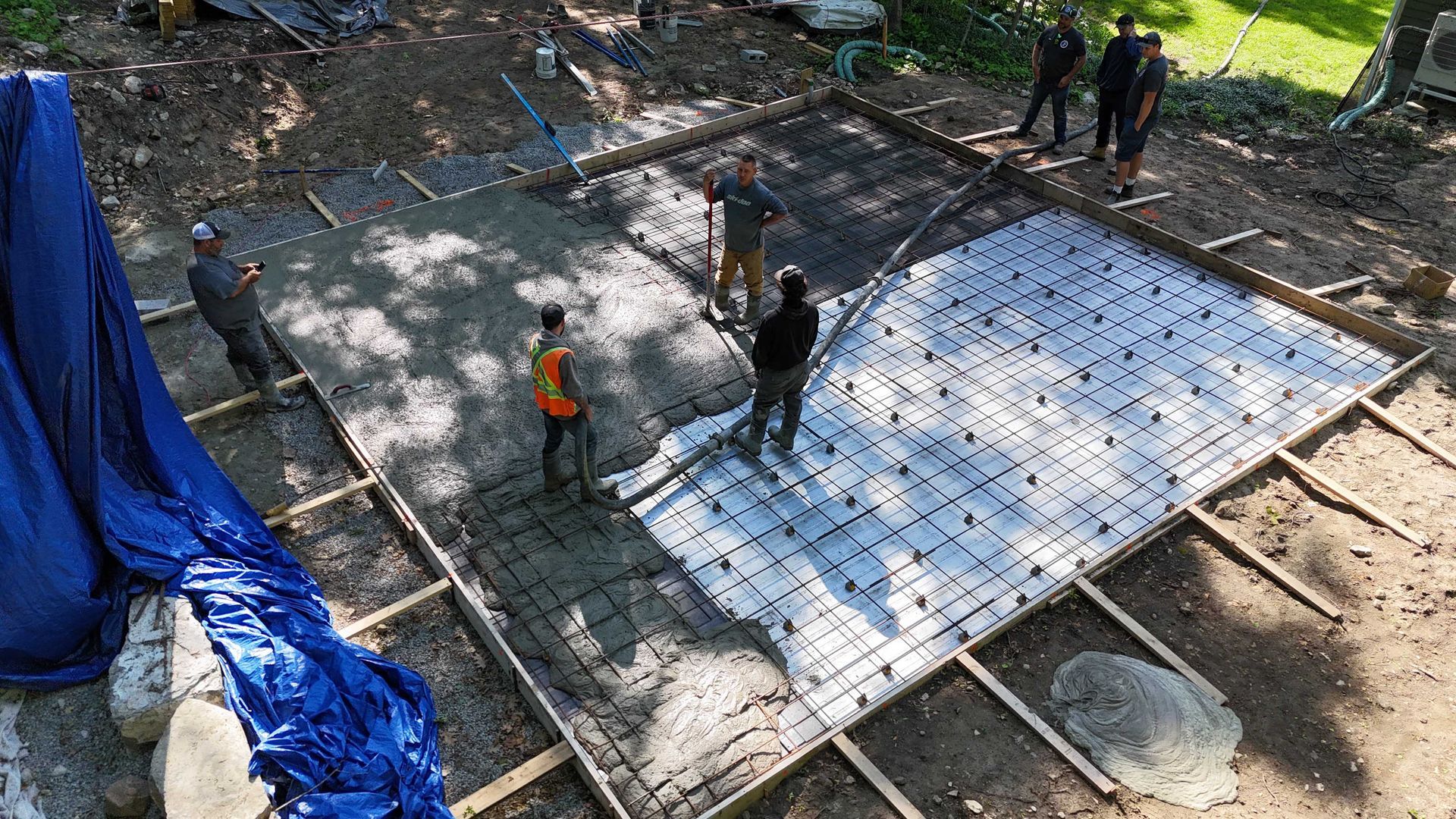 Workers pouring a concrete slab with rebar grid at a construction site, surrounded by blue tarp and dirt.