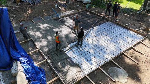 Workers pouring a concrete slab with rebar grid at a construction site, surrounded by blue tarp and dirt.