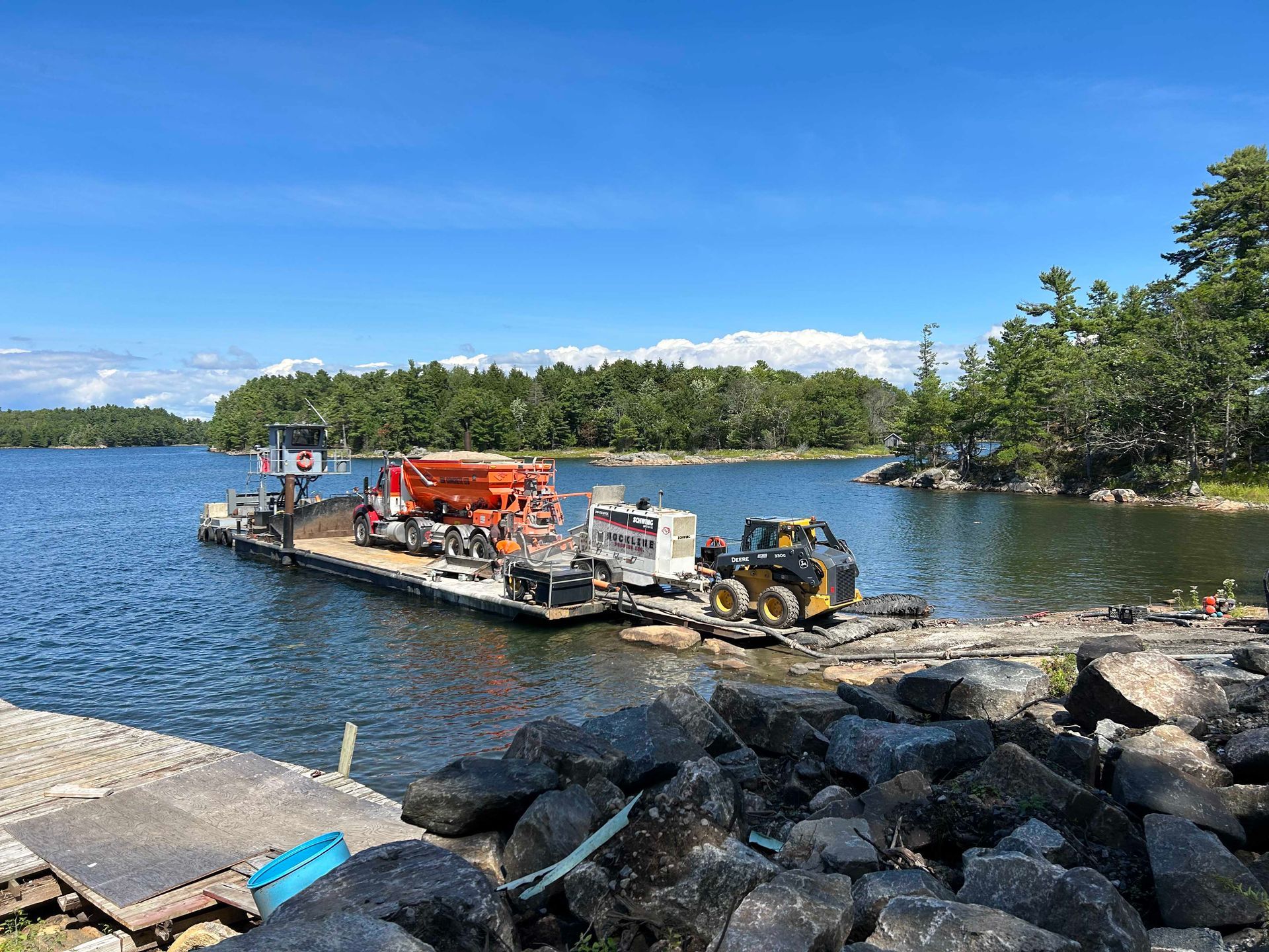 Construction barge with equipment working near a rocky shoreline on a lake under a blue sky
