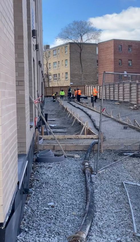 Construction workers on a building site with rebar, gravel, and wooden forms between brick buildings