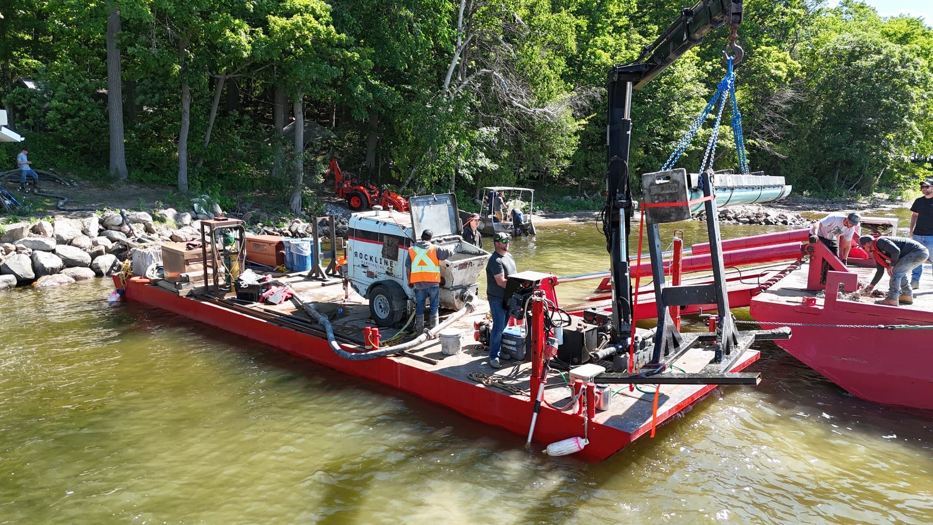 Red work barge with machinery and workers on a river beside a wooded shore