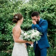 Bride and groom outdoors, exchanging vows; he wipes a tear while she holds a white bouquet.
