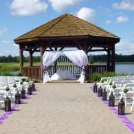 Outdoor wedding gazebo by a lake with white chairs, purple aisle accents, and draped arch
