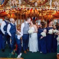 Wedding party posing on a carousel ride, holding white bouquets in front of lit carousel horses