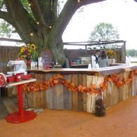 Rustic outdoor bar with autumn garland, red stool, and large tree in the background