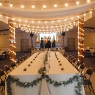 Decorated banquet hall with long white-draped tables, string lights, and a small stage at the back