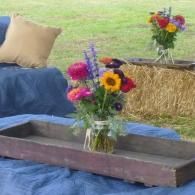 Flowers in glass vases on a blue-covered rustic table outdoors, with hay bales and a pillow in the background