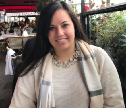 Smiling woman in a light sweater and scarf seated at an outdoor café, with plants and diners behind her