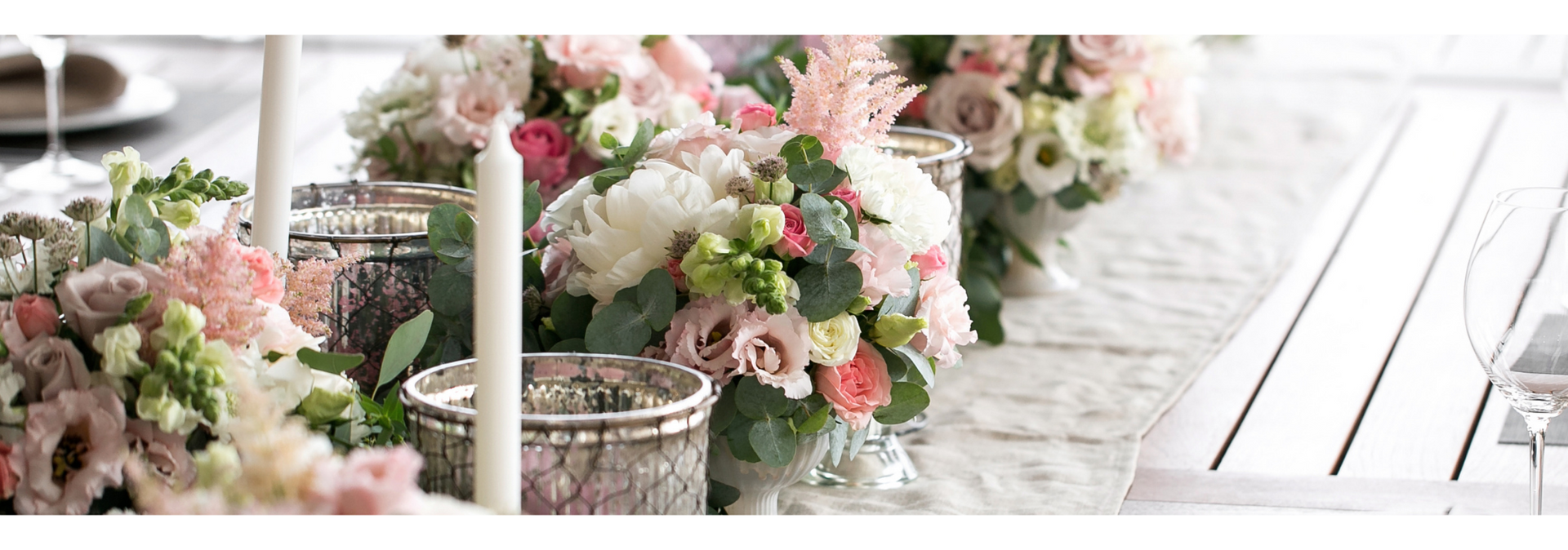 Pastel floral arrangements and candles line a white outdoor aisle beside a wooden deck