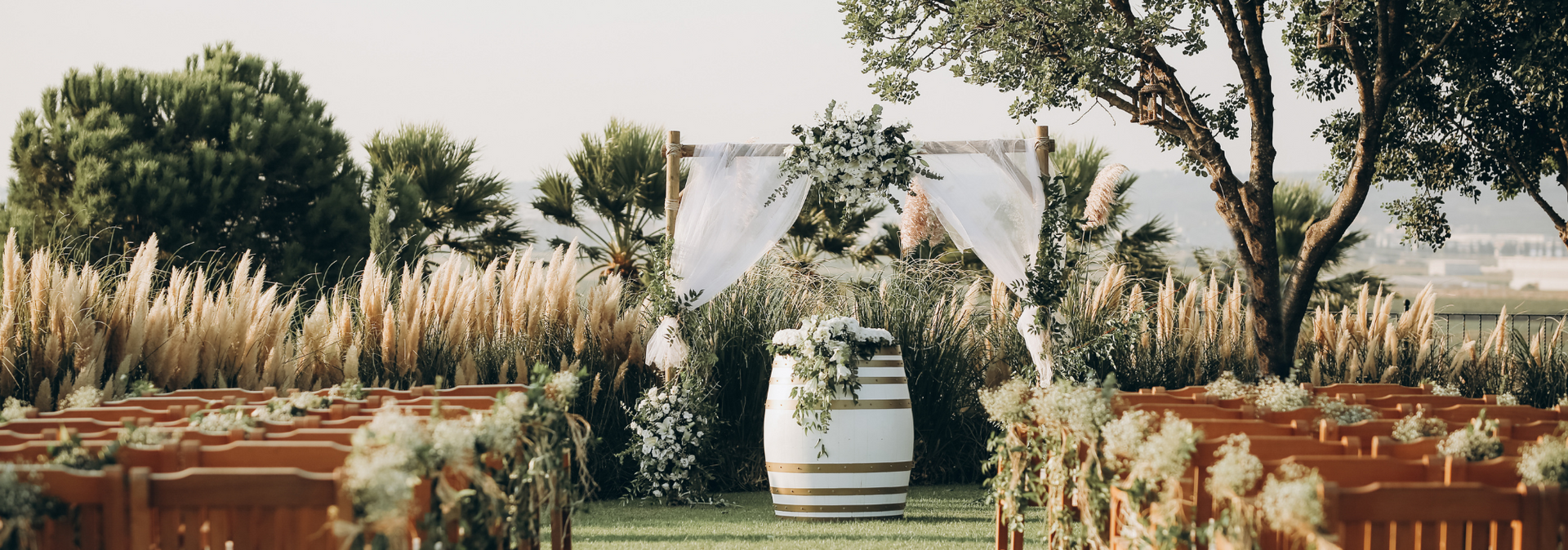 Outdoor wedding aisle with white flowers, rustic benches, and a decorated white barrel under trees