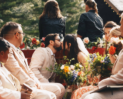 Guests seated outdoors at a flower-filled wedding ceremony, with a couple leaning in to kiss.