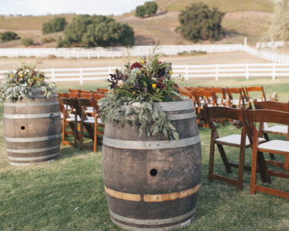 Rustic outdoor wedding setup with wooden barrels, floral arrangements, and rows of wooden chairs on a grassy lawn