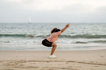 Person in pink top and black shorts squats on a sandy beach with arms extended, ocean behind.