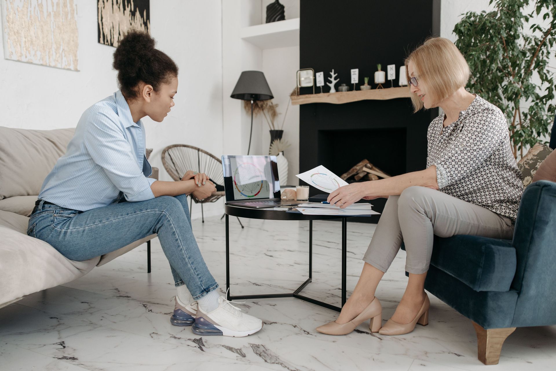 Two women seated in a living room discuss papers at a coffee table.