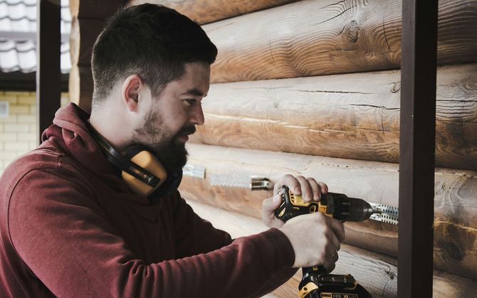 Man drilling into a log cabin wall beside a window, wearing a maroon hoodie and headphones around his neck