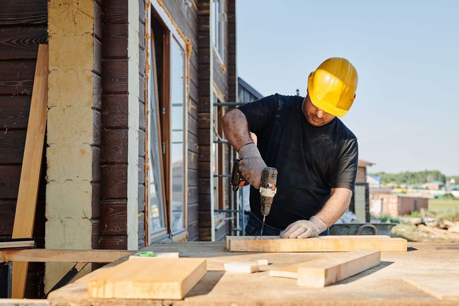 Worker in a yellow hard hat using a drill on wooden boards at a construction site