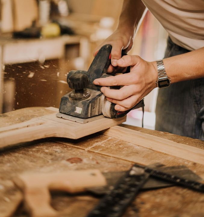 Person sanding a wooden board with a power sander in a woodworking shop, with sawdust flying