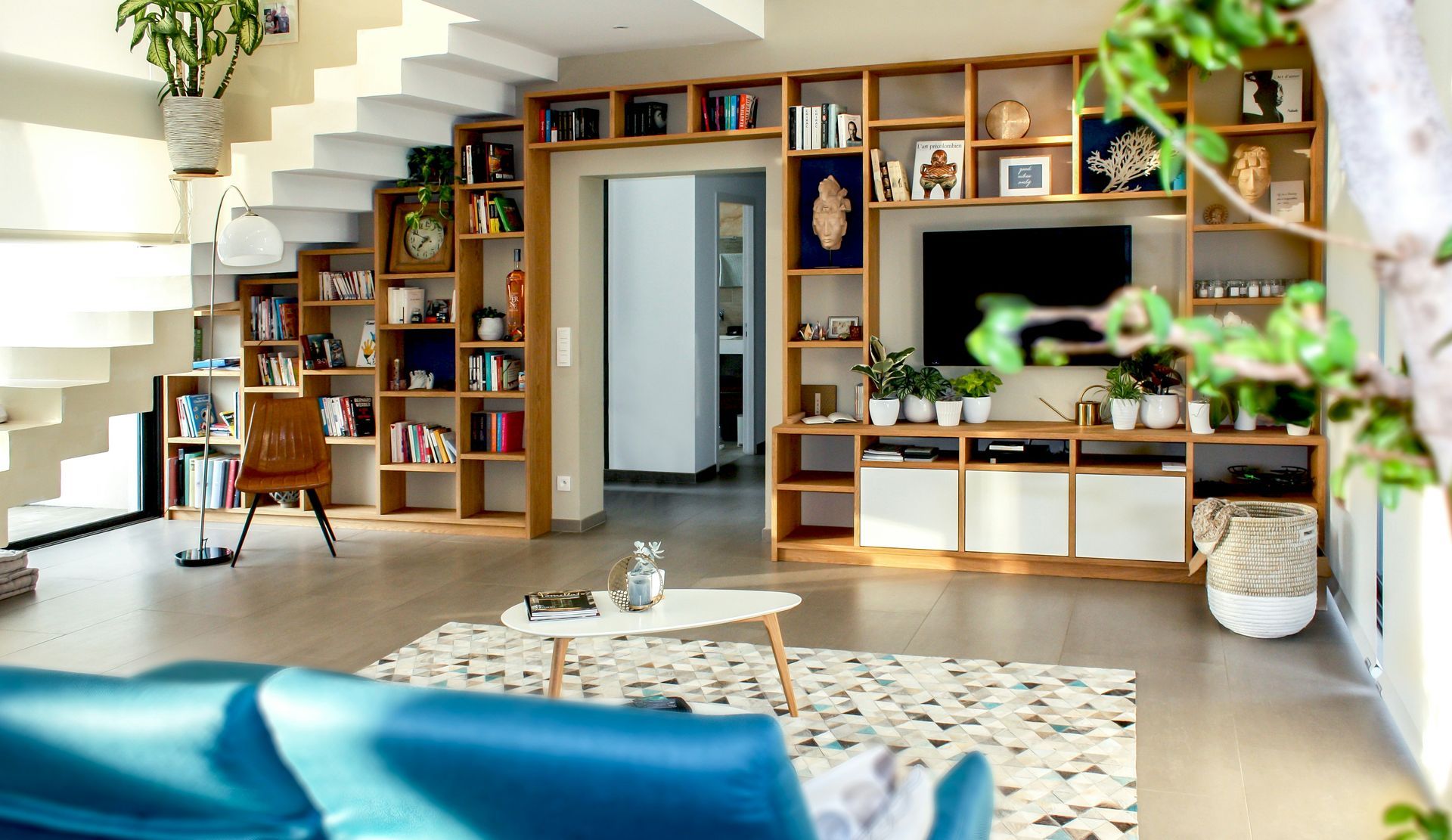 Bright modern living room with built-in shelves, wooden staircase, and blue sofa accents