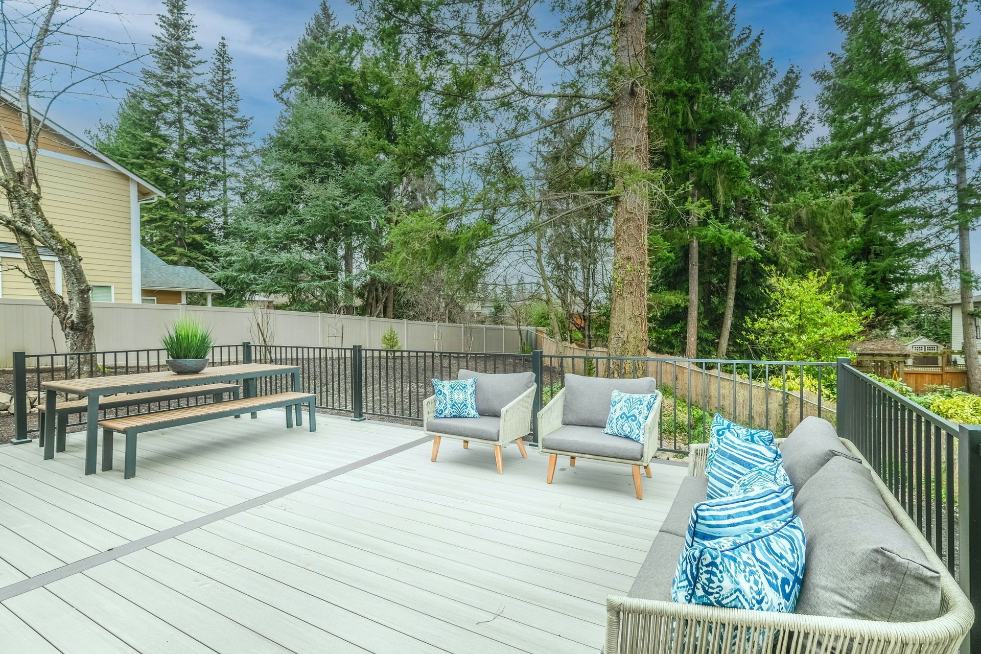 Spacious backyard deck with wicker seating, picnic table, and blue pillows, overlooking trees and a fenced yard